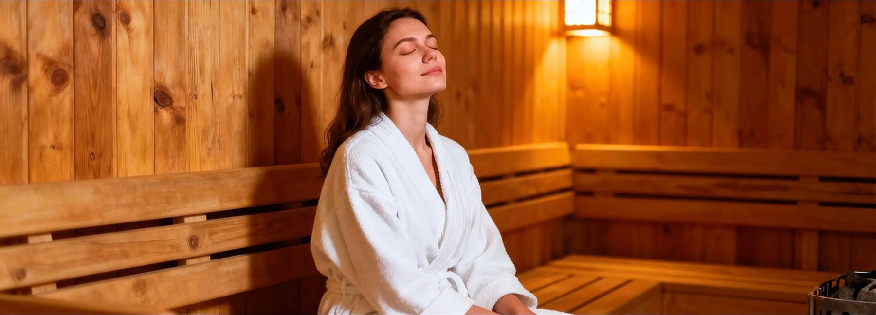 Woman in a white robe sitting in a wooden sauna with her eyes closed.
